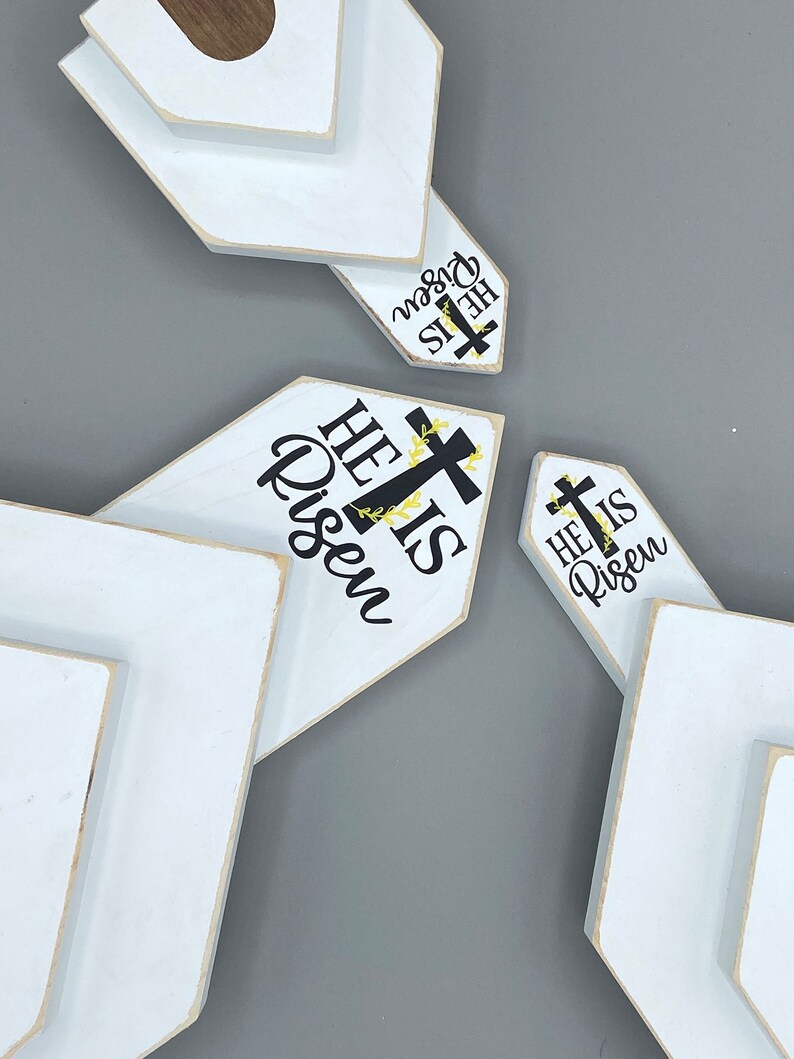 May include: Three white wooden signs with a chevron shape. Each sign has the text "He is Risen" with a black cross and yellow floral accents.