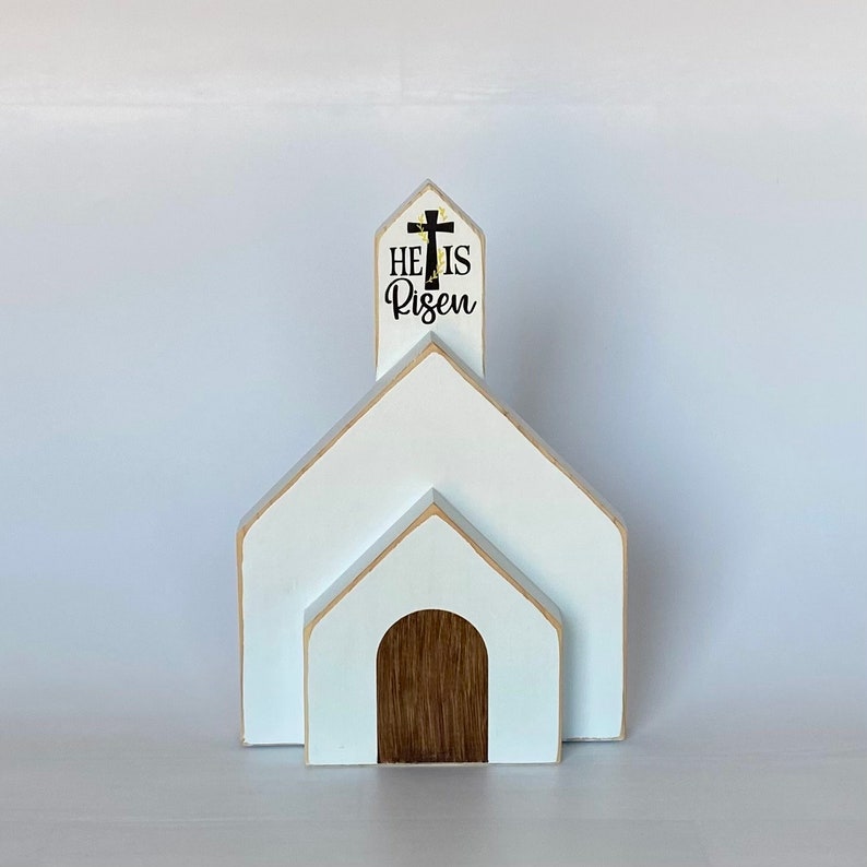May include: White wooden church with a brown door and a steeple that reads "He is Risen" with a black cross on top.