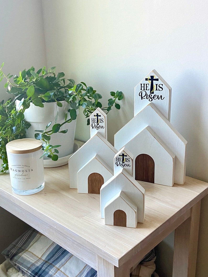 May include: Three white wooden church-shaped decorations with the text "He is Risen" and a cross on each. The decorations are arranged on a light wood table with a green plant in a white pot in the background.