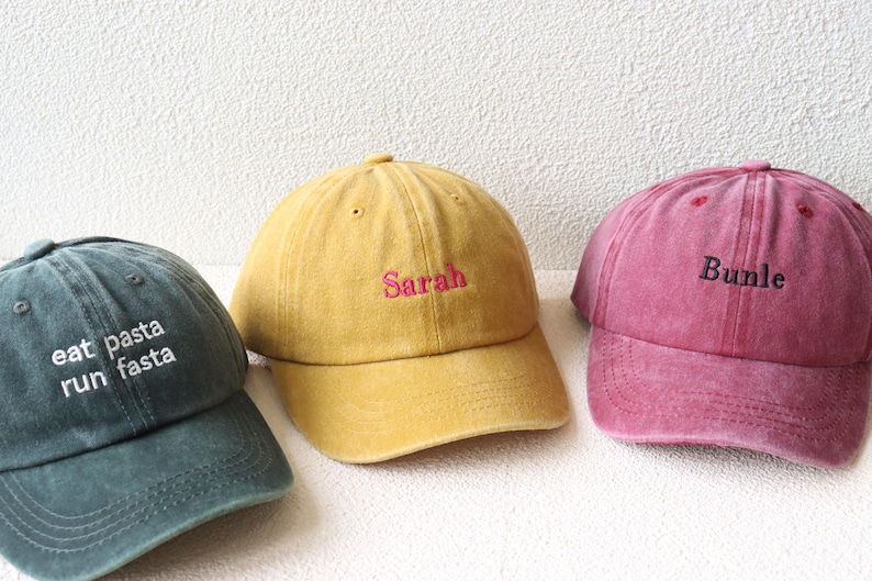 May include: Three different colored baseball caps with embroidered text. The first cap is green with white text that reads "eat pasta run fasta". The second cap is yellow with pink text that reads "Sarah". The third cap is pink with black text that reads "BUNDLE".