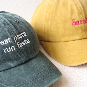 May include: Two baseball caps, one green with the text "eat pasta run fasta" embroidered in white, and one yellow with the name "Sarah" embroidered in pink.