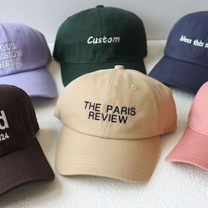 Könnte beinhalten: Sechs verschiedene Baseballkappen mit verschiedenen gestickten Texten. Die Kappen sind in verschiedenen Farben, darunter lila, grün, marineblau, braun, beige und rosa.