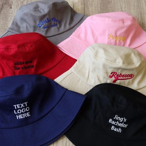 Könnte beinhalten: Eine Auswahl an bunten Fischerhüten in Rot, Rosa, Grau, Marineblau, Beige und Schwarz. Jeder Hut ist mit Text bestickt, darunter "Club One", "Poppy", "Rebecca", "Jing's Bachelor Bash", "embrace the chaos" und "TEXT LOGO HERE".