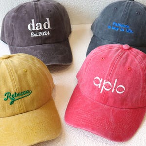 Könnte beinhalten: Vier Baseballkappen in verschiedenen Farben mit gesticktem Text. Die Kappen sind grau, rot, gelb und blau. Der Text auf den Kappen lautet "dad Est. 2024", "Patience is key in life", "Rebecca" und "aplo".