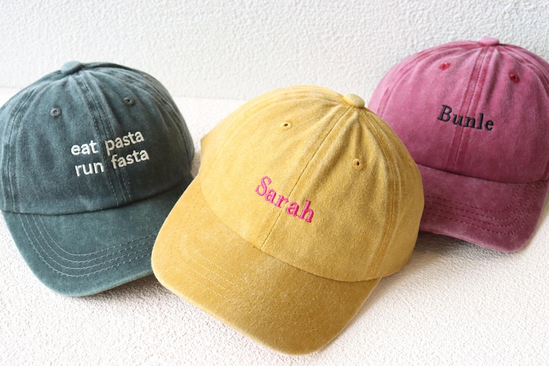 May include: Three different colored baseball caps with embroidered text. The first cap is dark green with white text that says "eat pasta run fasta". The second cap is yellow with pink text that says "Sarah". The third cap is pink with black text that says "Bundle".
