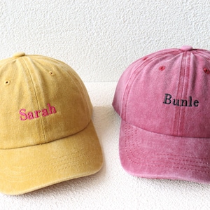 May include: Two baseball caps, one yellow with the name "Sarah" embroidered in pink and one pink with the name "Bundle" embroidered in black.