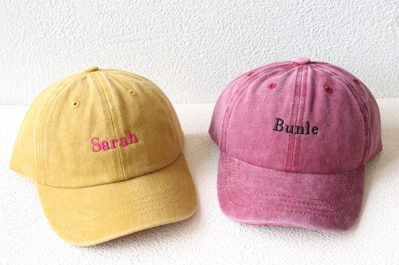 May include: Two baseball caps, one yellow with the name "Sarah" embroidered in pink and one pink with the name "Bundle" embroidered in black.