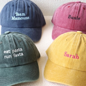 Könnte beinhalten: Vier Baseballkappen in verschiedenen Farben mit besticktem Text. Die Kappen sind blau, rot, grün und gelb. Der Text auf den Kappen lautet "Team Mamoune", "Bundle", "eat pasta run fasta" und "Sarah".