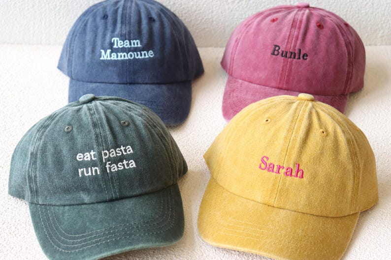 May include: Four different colored baseball caps with embroidered text. The caps are blue, red, green, and yellow. The text on the caps reads "Team Mamoune", "Bundle", "eat pasta run fasta", and "Sarah".
