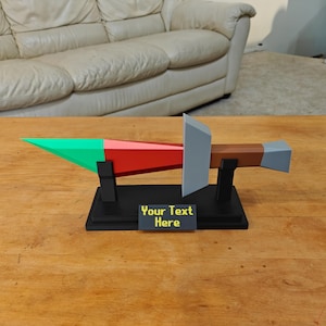 Pode incluir: Um modelo de espada geométrica com ponta verde, seção de lâmina vermelha, guarda cinza e cabo marrom com pomo cinza. A espada repousa sobre um suporte preto com uma placa retangular exibindo o texto amarelo Your Text Here.