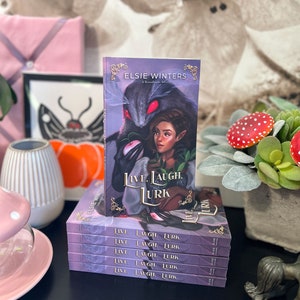 May include: A stack of five paperback books with a purple cover and gold lettering. The top book features a fantasy illustration of a creature with red eyes and a woman with long brown hair. The title of the book is "Live. Laugh. Lurk."