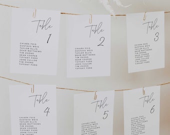 May include: Six white rectangular table seating cards hanging from a thin brown twine string with gold paper clips. Each card features the word Table followed by a number from one to six in black script, and a list of names in black sans-serif font.
