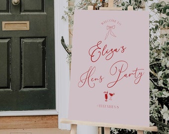 Puede incluir: Un letrero de bienvenida rectangular de color rosa claro sobre un caballete de madera con texto rojo que dice Welcome to Eliza's Hens Party, un dibujo lineal rojo de un lazo, dos ilustraciones de copas de cóctel y el hashtag #ELIZAHENS.