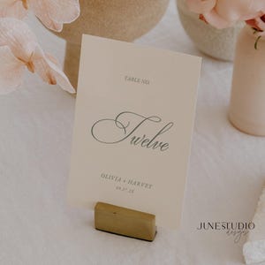 May include: A cream-colored table number card with the word "Twelve" in elegant script. The card also includes the names "Olivia + Harvey" and the date "09.27.2X". The card is held in a small, gold-colored wooden holder.