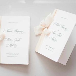 May include: Two white wedding programs with cream-colored ribbon and bows. The programs have elegant script text that reads "Natalia Montgomery and Martin Joseph Matthews" and the date "26th September 2020".