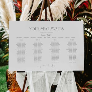 Peut inclure: Un panneau blanc avec le texte "YOUR SEAT AWAITS" et une liste de noms, probablement pour un mariage. Le panneau est sur un chevalet en bois, avec du tissu décoratif et de la verdure en arrière-plan. Le texte "bridal table" est également visible.