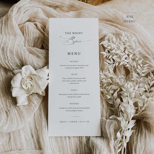 May include: A white menu card with the text "THE NIGHT Before MENU" in elegant script. The menu lists entree, main, sides, and dessert options. The card is placed on a beige fabric with a white flower and decorative sprigs.