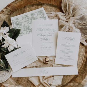 May include: Wedding stationery suite featuring calligraphy and floral patterns. The set includes an invitation, RSVP card, details card, and a belly band. The items are arranged on a wooden tray with fabric and flowers.