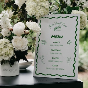Könnte beinhalten: Eine weiße Speisekarte mit grünem Rand und dem Text "Taco 'bout love" und "MENU". Die Speisekarte listet Fleischsorten, Beläge, Saucen und Beilagen auf. Die Speisekarte ist von weißen Blumen und Grün umgeben.