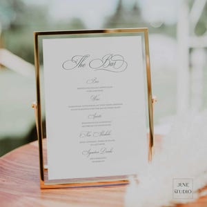 May include: A gold-framed sign with a white background and black text lists the bar menu for a wedding. The menu includes beer, wine, spirits, non-alcoholic drinks, and signature drinks.