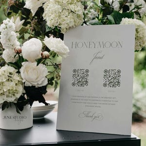 May include: White sign with the text "Honeymoon Fund" and two QR codes. The sign is on a black surface with white flowers in a vase.