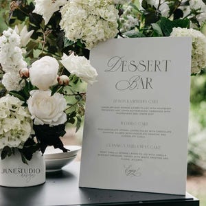 May include: A white sign with black text listing dessert options for a wedding reception. The sign reads "Dessert Bar", "Lemon Raspberry Cake", "Wedding Cake", and "Classic Vanilla Bean Cake".