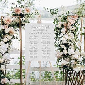 Könnte beinhalten: Ein weißer Hochzeitssitzplan mit dem Text "Your seat awaits" und den Namen Eliza & Harvey. Der Plan steht auf einem Staffelei mit Blumenarrangements auf beiden Seiten.
