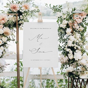 May include: A wedding welcome sign on a wooden easel, with the names Mia & Simon in elegant script. The sign is surrounded by floral arrangements of white and peach flowers, with a backdrop of greenery and a lake.