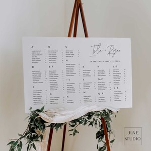 May include: A white rectangular wedding seating chart sign with black text listing guest names alphabetically by letter, featuring the names Isla + Ryan and the date 16 September 2023, displayed on a wooden tripod easel with draped fabric and greenery.