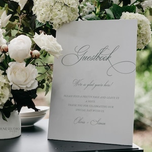 Könnte beinhalten: Ein weißes Gästebuchschild mit eleganter Schrift, auf dem "Guestbook" steht. Das Schild enthält den Text "Wir freuen uns so sehr, dass Sie hier sind!" und eine Nachricht, um eine Notiz zu hinterlassen. Weiße Blumen und Grünzeug sind im Hintergrund.
