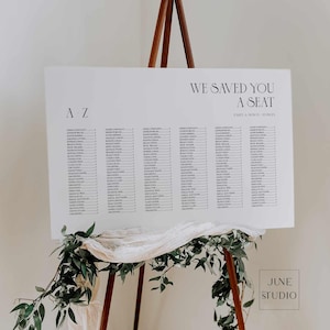Op de afbeelding: Een witte plattegrond met de tekst "WE SAVED YOU A SEAT" en een lijst met namen. De kaart wordt weergegeven op een houten ezel, versierd met groen en stof. De tekst "A - Z" staat in de linkerbovenhoek.