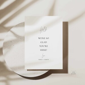 May include: A white card with the text "Hello, We're so glad you're here!" and the names "Emily + Simon" at the bottom. The card is on a light brown surface with shadows from a plant.