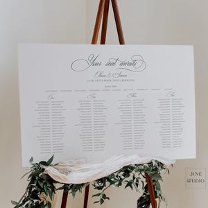 May include: A white rectangular wedding seating chart sign with black text reading Your seat awaits, Olivia & Simon, 14th September 2025, Burrimo, and a list of guest names organized by table numbers, displayed on a wooden easel with green foliage.