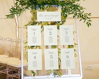 May include: A clear acrylic wedding seating chart sign on a white easel, decorated with green foliage. The sign features the text Our favourite people, Emily & Simon, and lists guest names under six numbered table cards in black serif font.