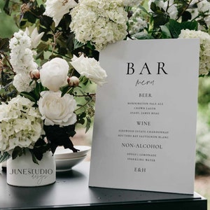 May include: A white sign with black text listing a bar menu with beer, wine, and non-alcoholic options. The sign is on a black table with a white vase of white flowers in front of it.