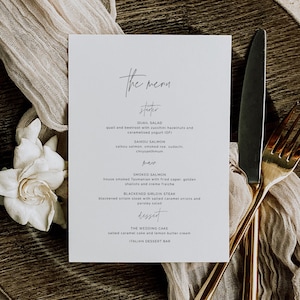 May include: A white menu card with elegant calligraphy, listing a wedding menu. The menu includes starters, main courses, and dessert options. A gold fork and knife are placed on the right side of the card, with a white flower and fabric in the background.