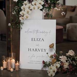 May include: A wedding welcome sign with the names ELIZA and HARVEY, surrounded by floral arrangements and candles. The sign reads "Welcome to the wedding of Eliza and Harvey, so glad you're here!" with the date and location.