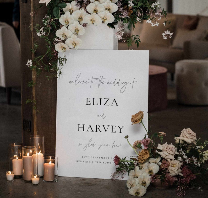 May include: A wedding welcome sign with the names ELIZA and HARVEY, surrounded by floral arrangements and candles. The sign reads "Welcome to the wedding of Eliza and Harvey, so glad you're here!" with the date and location.