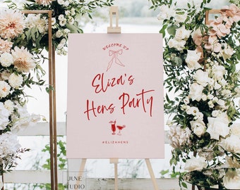 Könnte beinhalten: Ein rechteckiges, hellrosa Willkommensschild auf einer Holzstaffelei mit rotem Text, der Welcome to Eliza's Hens Party lautet, einer roten Strichzeichnung einer Schleife, zwei Cocktailgläsern und dem Hashtag #ELIZAHENS unten.