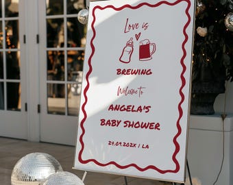 May include: A rectangular white sign with a wavy red border featuring red text and graphics. The sign reads Love is BREWING Welcome to ANGELA'S BABY SHOWER 24.09.202X / LA, with illustrations of a baby bottle and a beer mug separated by a small heart.