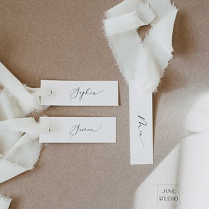 May include: Three white rectangular place cards with black calligraphy writing. The names on the cards are Sophia, Simon, and Mia. The cards are on a light brown surface with white fabric ribbons.