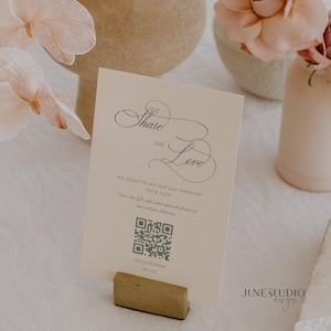 May include: A rectangular cream-colored card featuring the text Share the Love, We want to see our day through your eyes, Scan the QR code and upload photos to our online album, Olivia & Simon, and 08.17.25, held by a small gold metal stand.