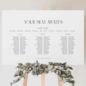 Peut inclure: Un plan de table de mariage blanc avec un design minimaliste. Le plan liste les noms des invités sous les titres "Un", "Deux" et "Trois". Le texte "Your Seat Awaits" est en haut du plan, avec les mots "bridal table" en dessous. Le bas du plan comprend le texte "Tyler & Simon, 23.08.2015".