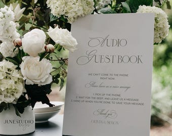 May include: A white sign with the text "Audio Guest Book" and instructions for leaving a voice message. The sign is surrounded by white flowers in a vase with the text "JUNE STUDIO designs".