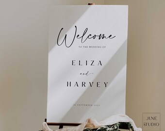 May include: A white rectangular wedding welcome sign featuring black text that reads Welcome to the wedding of Eliza and Harvey 16 September 2023. The sign is displayed on a metal easel decorated with green foliage and a beige fabric drape.
