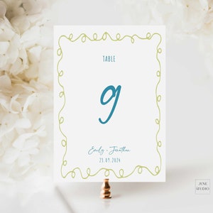 May include: A white table number card with a green curly border. The number 9 is written in blue. The text "Table" is written in black. The text "Emily & Jonathan" and "23.09.2024" is written in black.