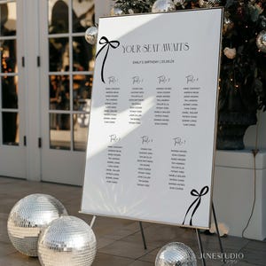May include: A white seating chart with black text and a gold frame, featuring guest names organized by table. The sign reads "YOUR SEAT AWAITS" and "EMILY'S BIRTHDAY". Decorative black ribbons adorn the top and bottom. Silver disco balls are in the foreground.