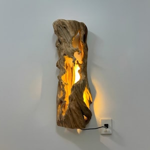 May include: A wooden wall sconce with a natural, organic shape. The sconce is made from light brown wood with a carved-out centre that emits a warm, yellow light. The sconce is plugged into a wall outlet.