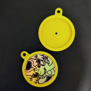 Puede incluir: Dos colgantes circulares amarillos con un pequeño lazo en la parte superior. Un colgante presenta un gráfico colorido de una criatura de dibujos animados. El otro colgante es amarillo liso con un pequeño agujero en el centro.
