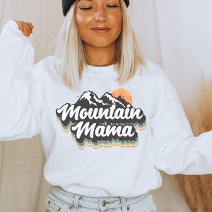 Könnte beinhalten: Weißer Pullover mit einer Retro-Grafik von Bergen und einer Sonne mit dem Text "Mountain Mama" in schwarzen und goldenen Buchstaben.